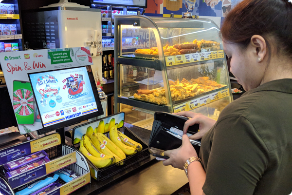 A yound women wearing a green shirt. reaches into her wallet at the counter of a conveinence store. The counter has power bars, bananas and an assortment of fried food.